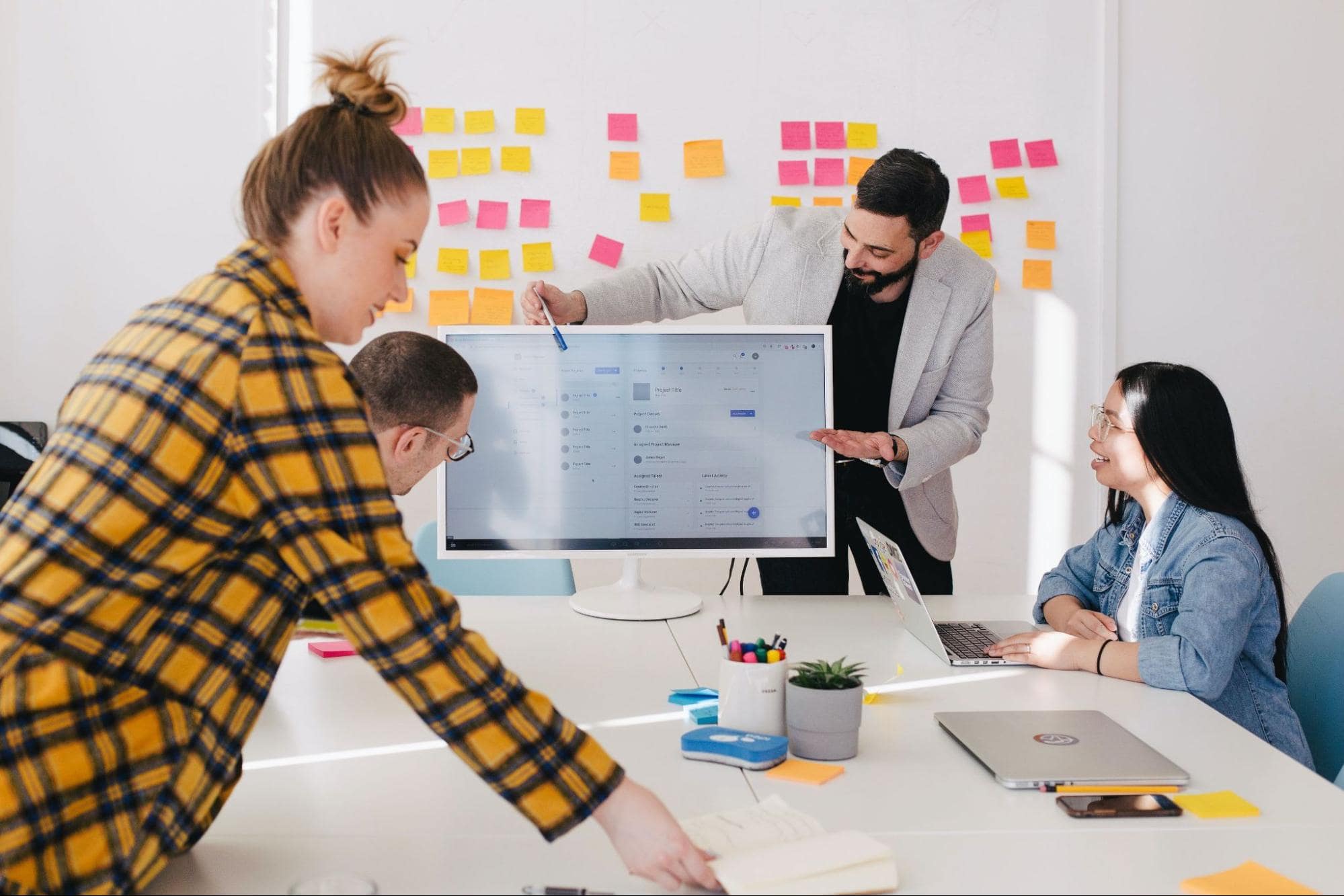 Team works around a large monitor displaying a project UI; man in a blazer points as sticky notes fill the wall.
