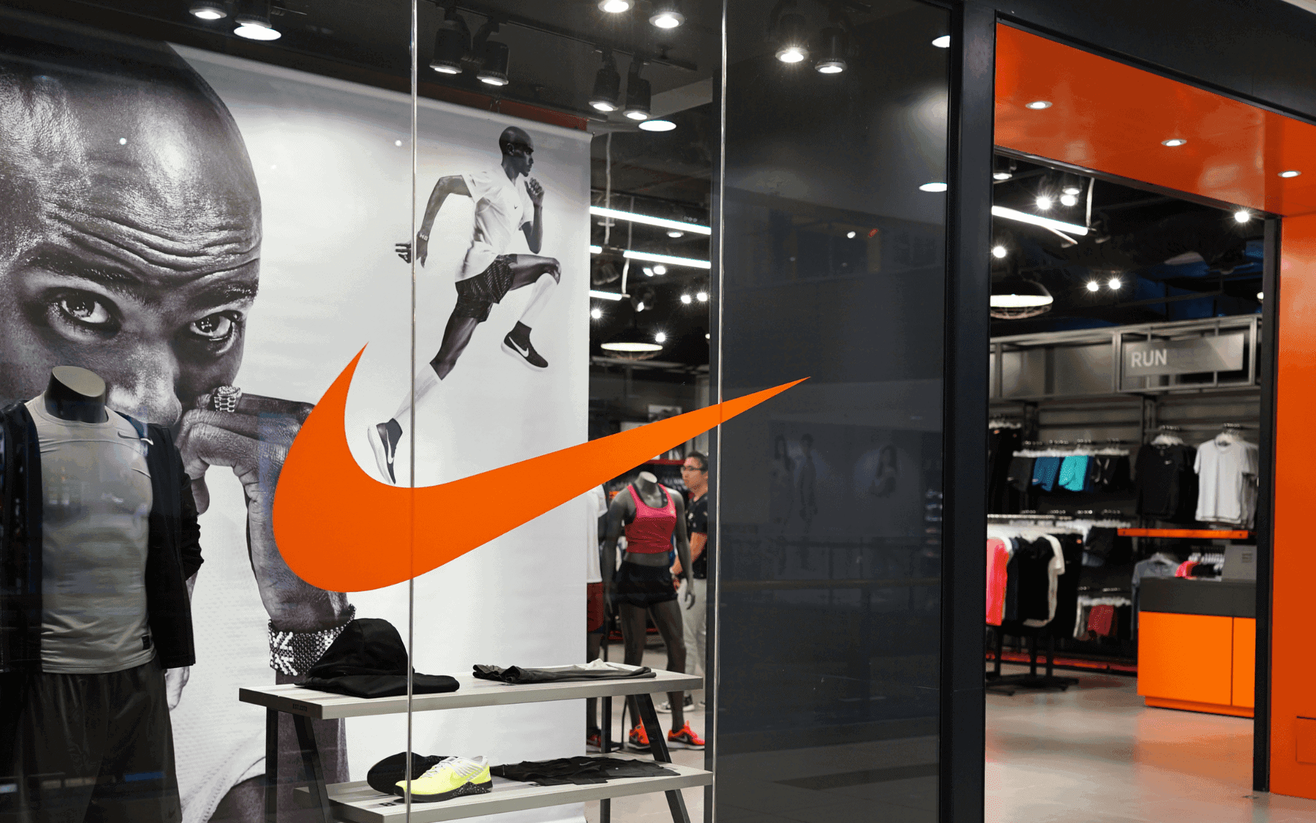 Nike storefront with giant orange swoosh logo, black-and-white athlete mural, mannequins, clothing racks, and a RUN sign.