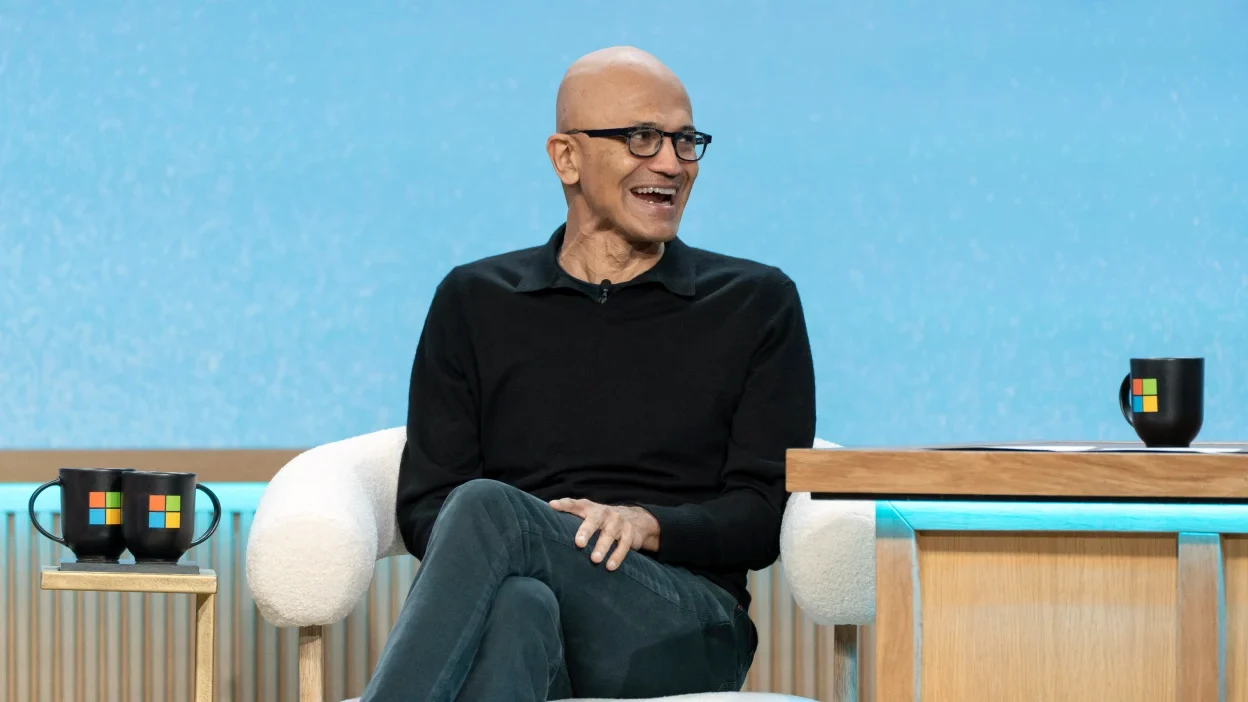 Speaker in black long-sleeve, glasses, bald, seated on a white chair in a tech-talk set with a blue backdrop.