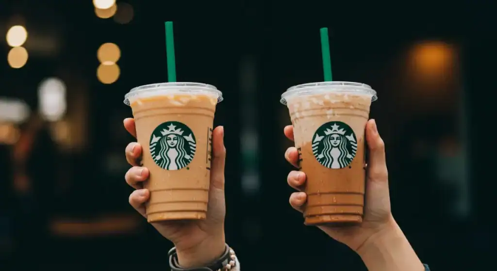 Two iced Starbucks drinks with green straws held up by hands, visible Starbucks logos on the cups.