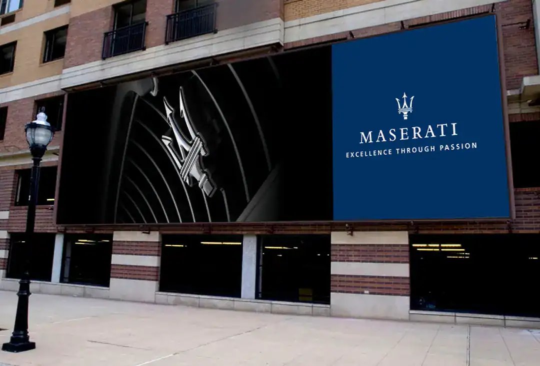 Maserati MASERATI EXCELLENCE THROUGH PASSION on a blue sign beside a chrome trident emblem against a black curved backdrop.