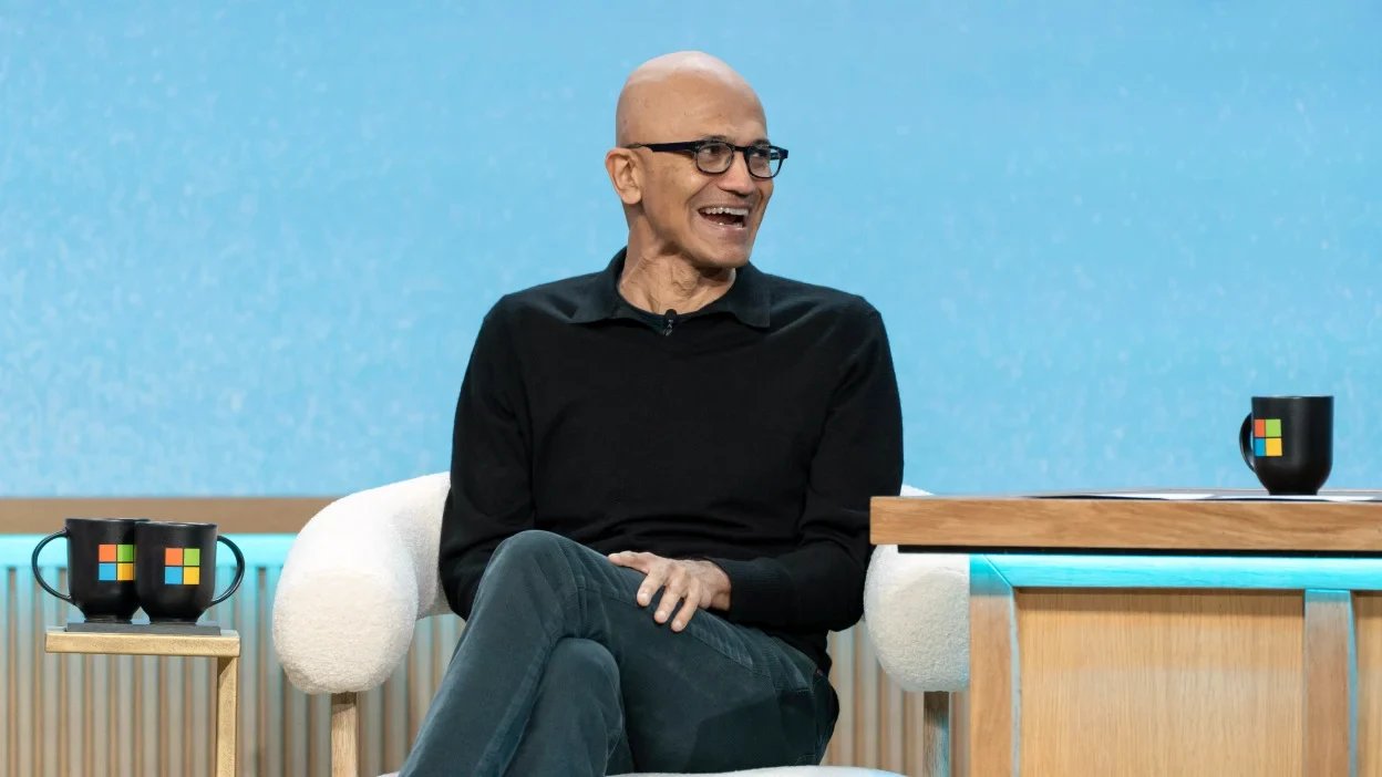 Speaker in black long-sleeve, glasses, bald, seated on a white chair in a tech-talk set with a blue backdrop.