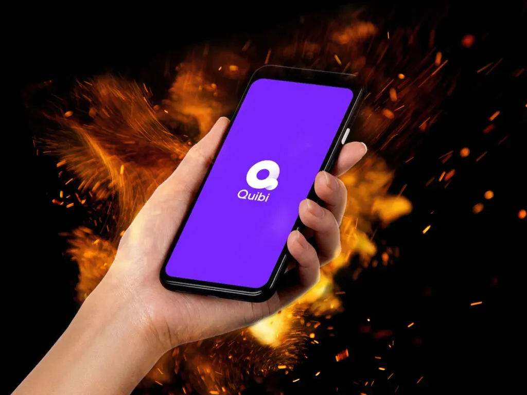 Hand Holding A Smartphone With A Purple Screen Displaying The Quibi Logo, Set Against A Dark Background With Orange Sparks.