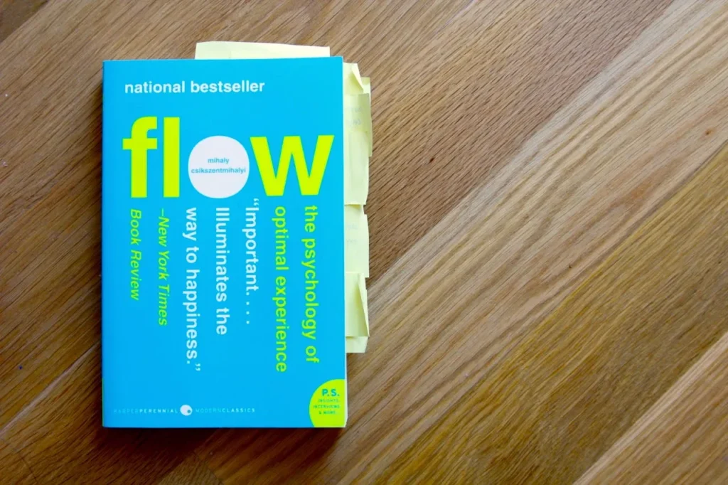 Flow: The Psychology Of Optimal Experience Book Cover On Blue, With Yellow-Green Typography And Sticky Notes On A Wooden Table.
