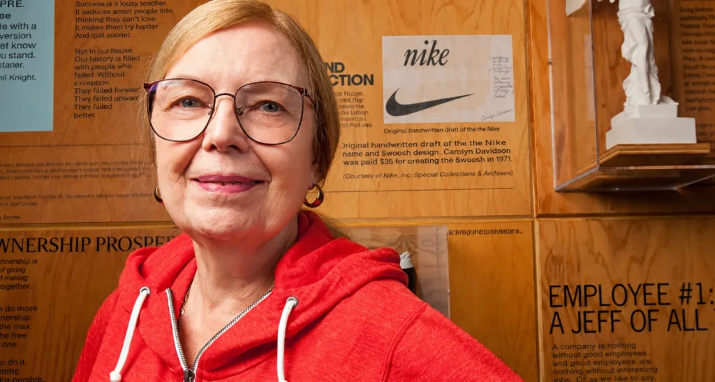 Portrait Of A Smiling Woman With Glasses In A Red Hoodie, Wooden Wall Display With Nike Sketch And Text Panels In The Background.